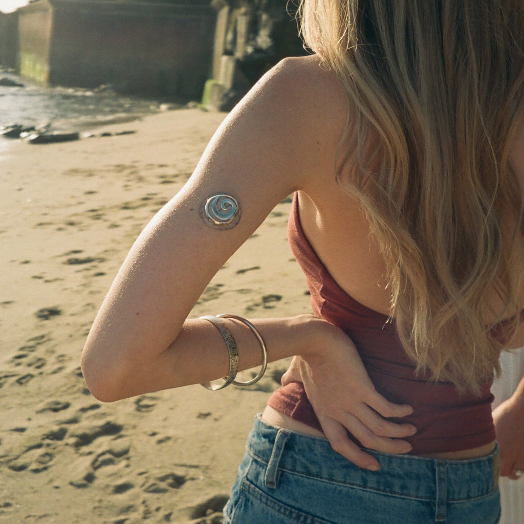 A woman on a sandy beach seen from behind, wearing a metallic circular temporary tattoo on her upper arm, a rust-colored top, and blue jeans.