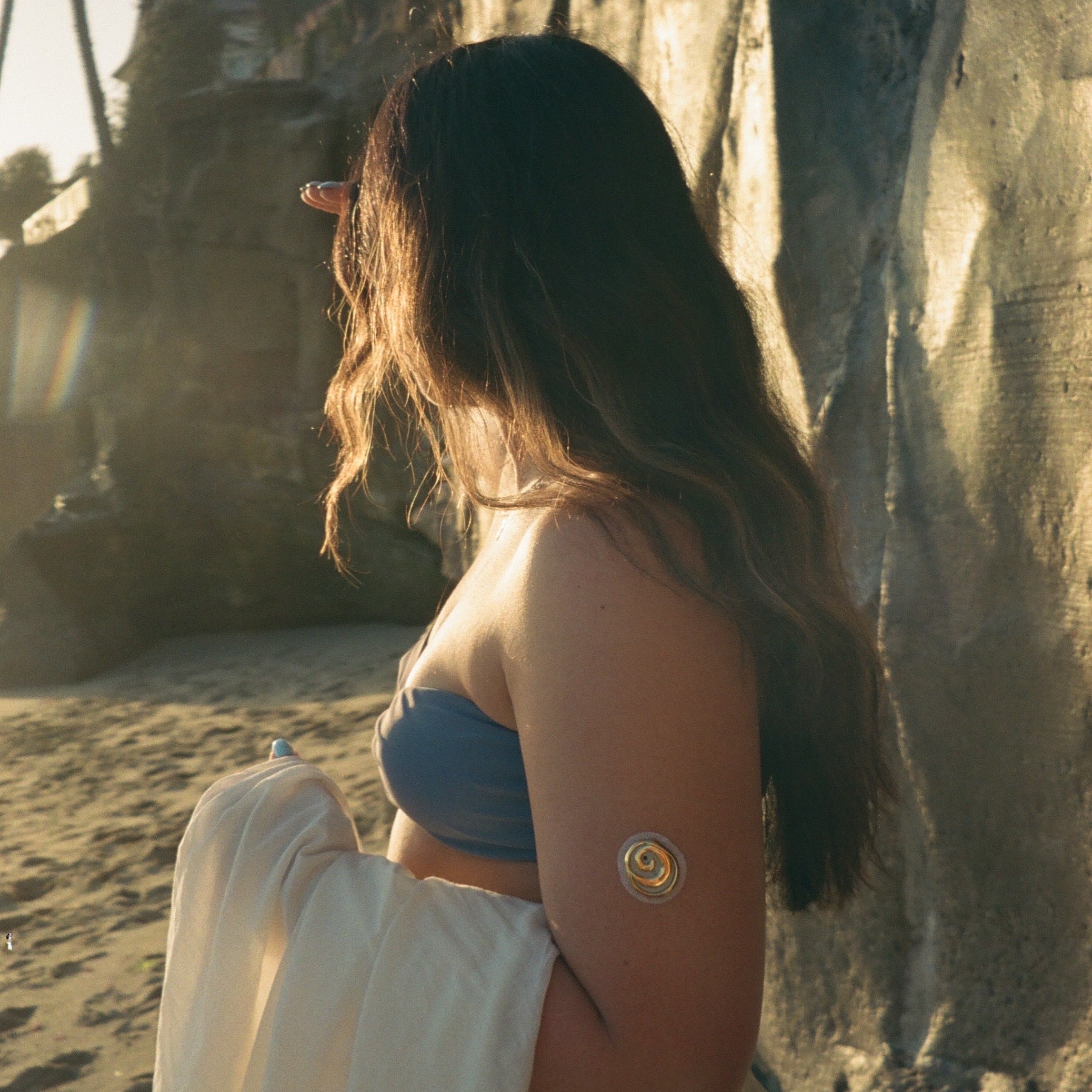 A woman with long dark hair stands on a sandy beach in profile, wearing a blue bandeau top and a flowing white skirt or drape, with a small circular gold-toned accessory on her upper arm. Sunlight is warm and directional, casting shadows on a large rock face behind her.