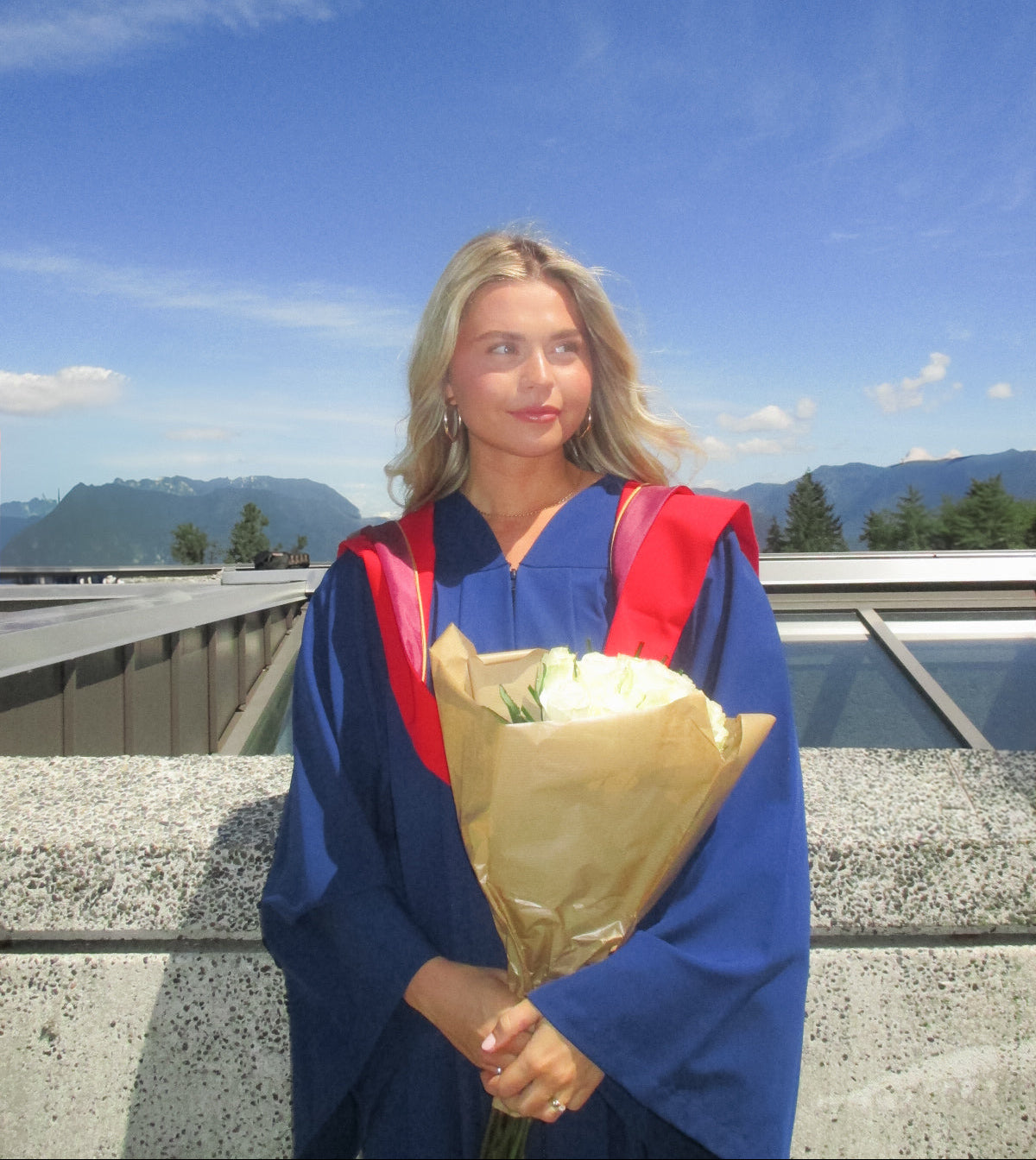 Graduate in blue gown with red hood holding flowers against a clear blue sky.