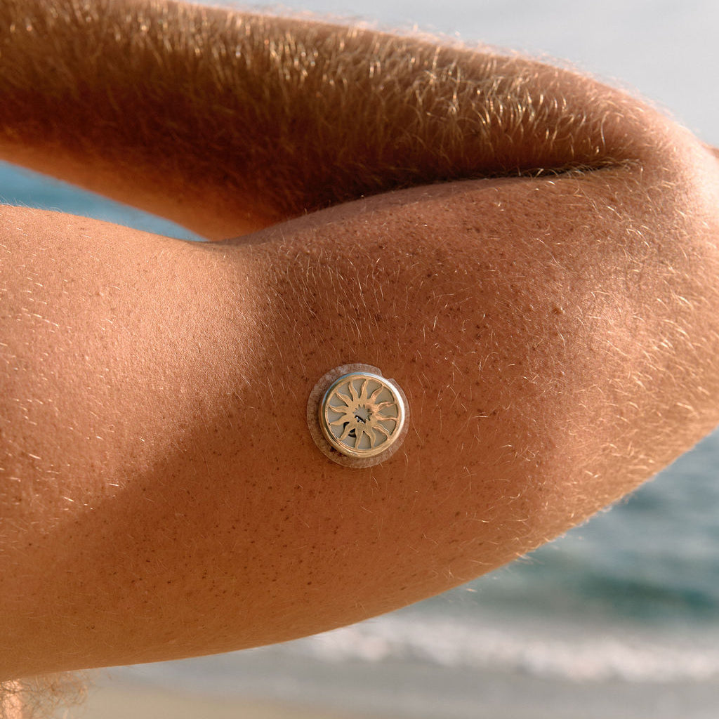 A close-up of a person's forearm at a beach, wearing a small round metallic charm or button with a sunburst design.